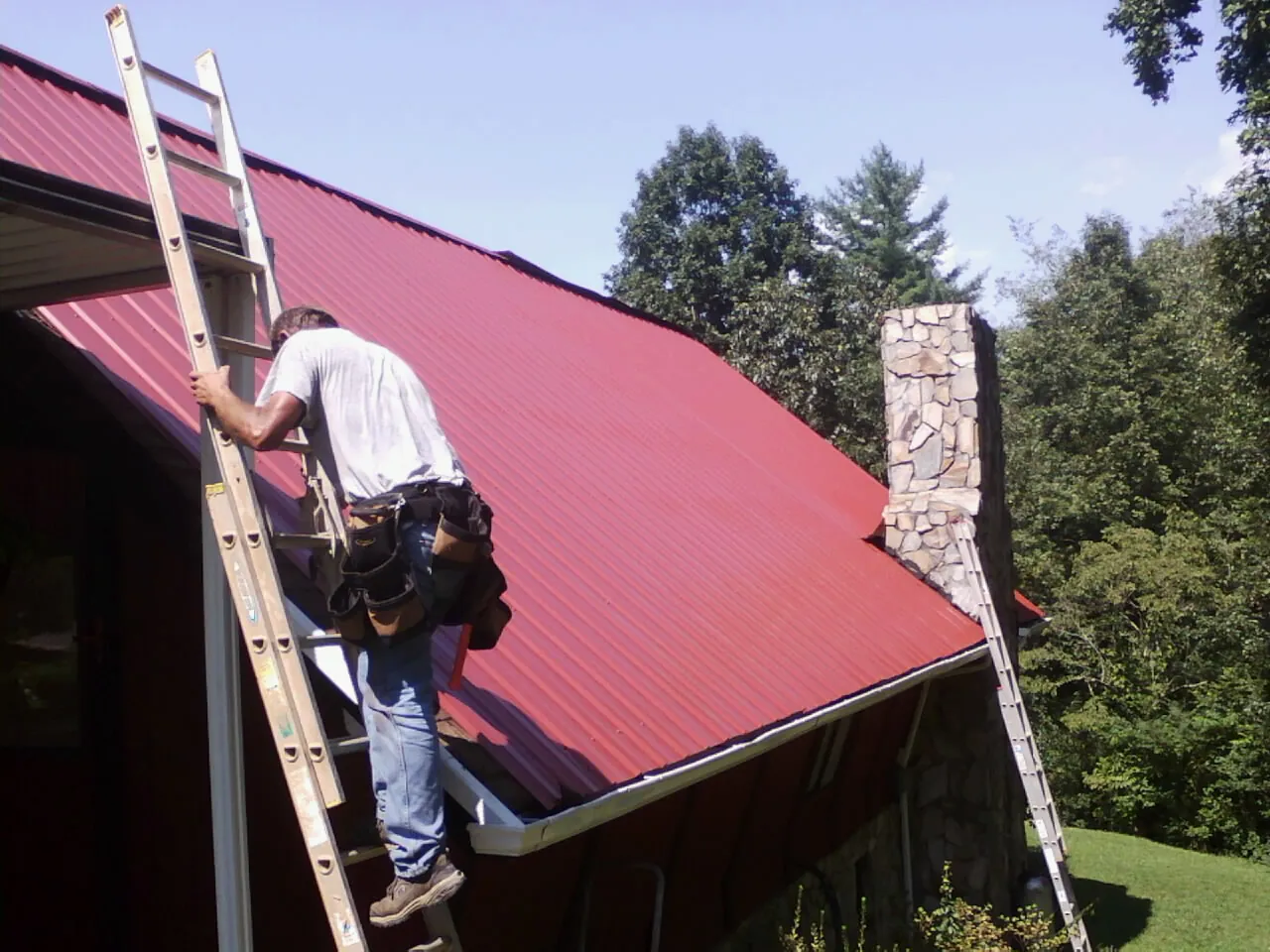 Roofer on a ladder beside a red metal roof and stone chimney — Soffit & Fascia Repair by Roofing Brothers in Youngwood