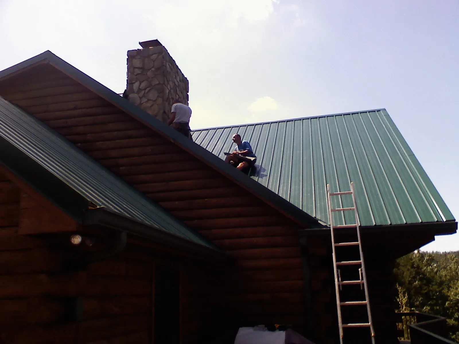 Roofers installing green metal panels beside a stone chimney on a log home in Youngwood